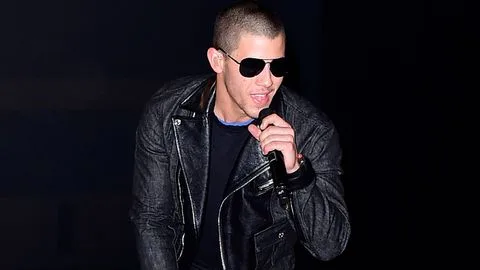 Ο Nick Jonas με μαύρο δερμάτινο μπουφάν και γυαλιά ηλίου τραγουδάει σε μικρόφωνο.