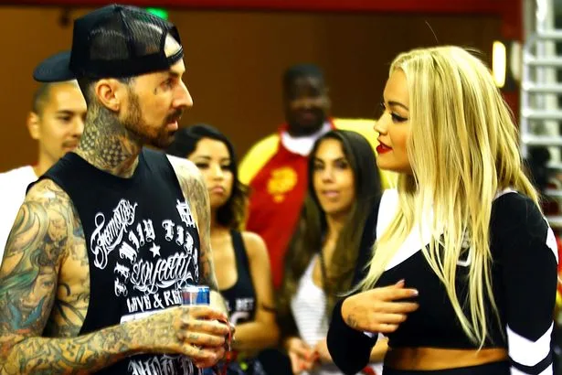 Ο Travis Barker και η Rita Ora συνομιλούν σε εσωτερικό χώρο.
