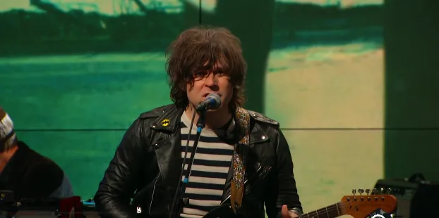 Ο Ryan Adams τραγουδάει σε σκηνή με κιθάρα και δερμάτινο μπουφάν.