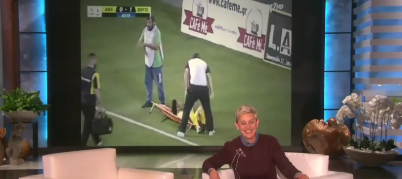 Η Ellen DeGeneres στο πλατό της εκπομπής της με φόντο βίντεο από ποδοσφαιρικό αγώνα.