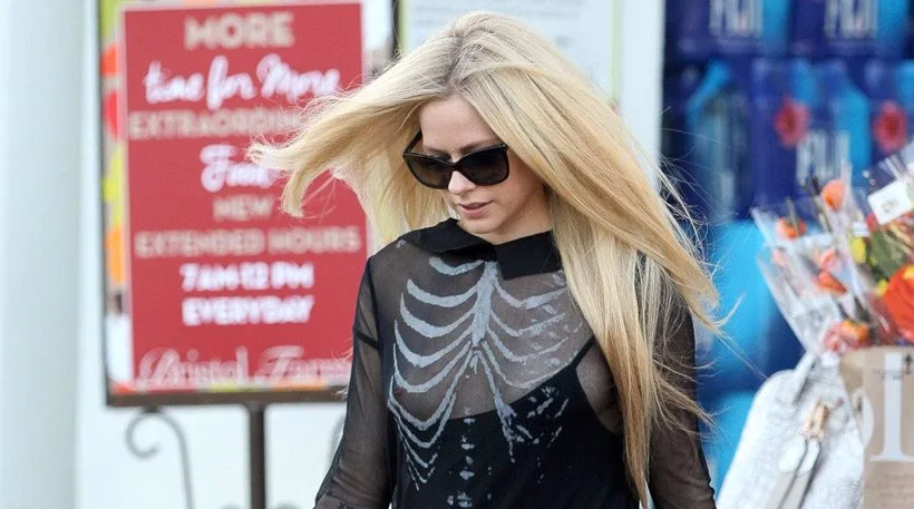 Η Avril Lavigne περπατά στο δρόμο φορώντας μαύρη μπλούζα με σχέδιο σκελετού και γυαλιά ηλίου.