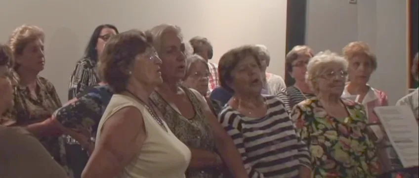 Μια ομάδα ηλικιωμένων γυναικών από τα ΚΑΠΗ Ηλιούπολης τραγουδούν σε χορωδία.