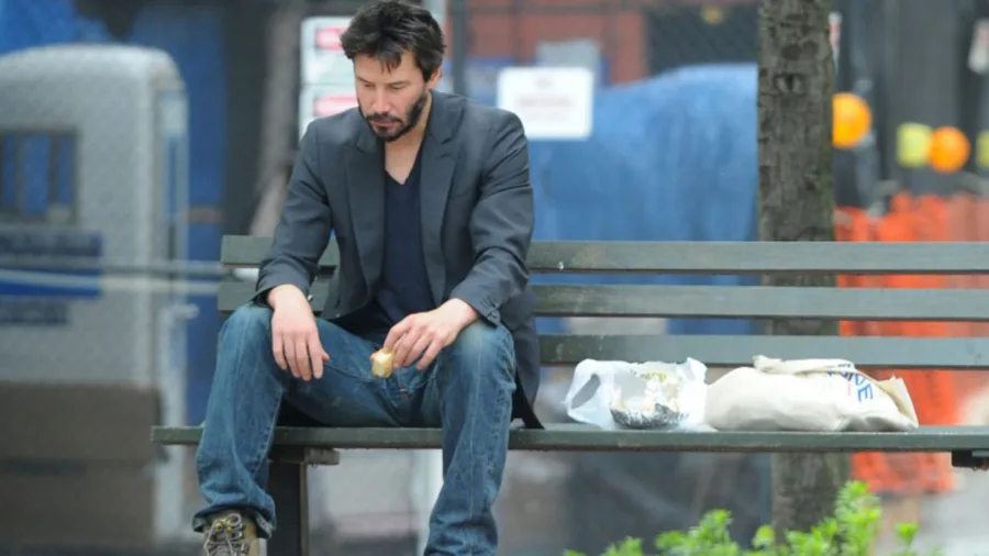 Ο Keanu Reeves κάθεται μόνος σε παγκάκι και τρώει.