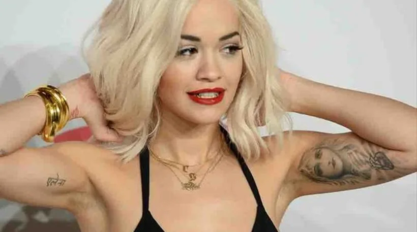 Η Rita Ora με ξανθά μαλλιά και κόκκινο κραγιόν ποζάρει με τα χέρια στο κεφάλι.