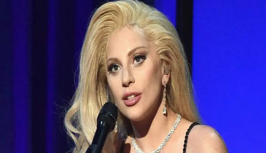 Η Lady Gaga με ξανθά μαλλιά και μαύρο φόρεμα μπροστά από μπλε φόντο