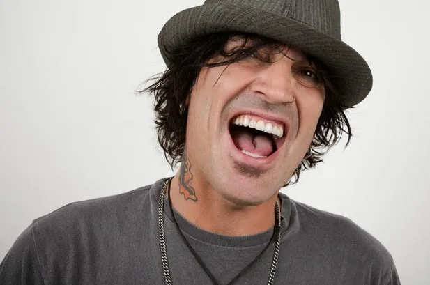 Ο Tommy Lee με καπέλο και γκρίζα μπλούζα γελάει.