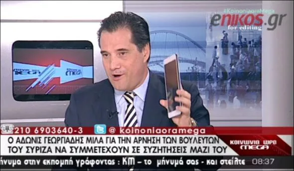 Άνδρας με κοστούμι σε τηλεοπτικό πάνελ κρατάει κινητό τηλέφωνο.