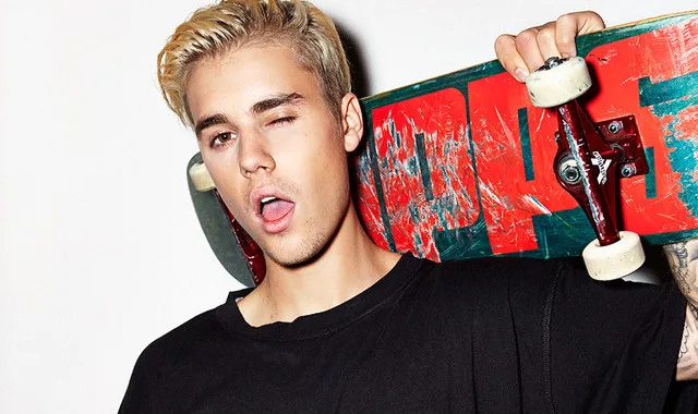 Ο Justin Bieber με ξανθά μαλλιά κρατάει ένα skateboard στον ώμο του.