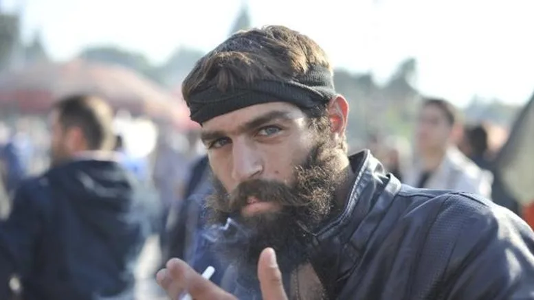Ο Στάθης Στιβακτάκης με μαύρο κεφαλόδεσμο και μούσια καπνίζει σε εξωτερικό χώρο.