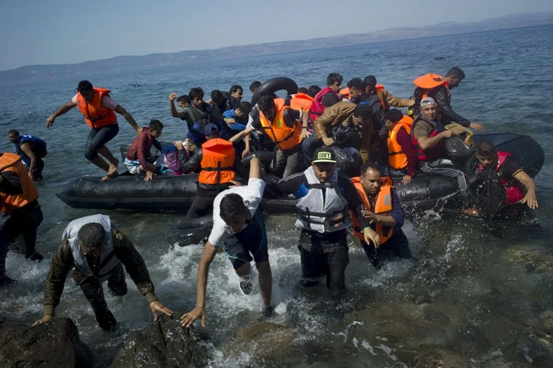 Πρόσφυγες και μετανάστες αποβιβάζονται από φουσκωτή βάρκα σε βραχώδη ακτή.