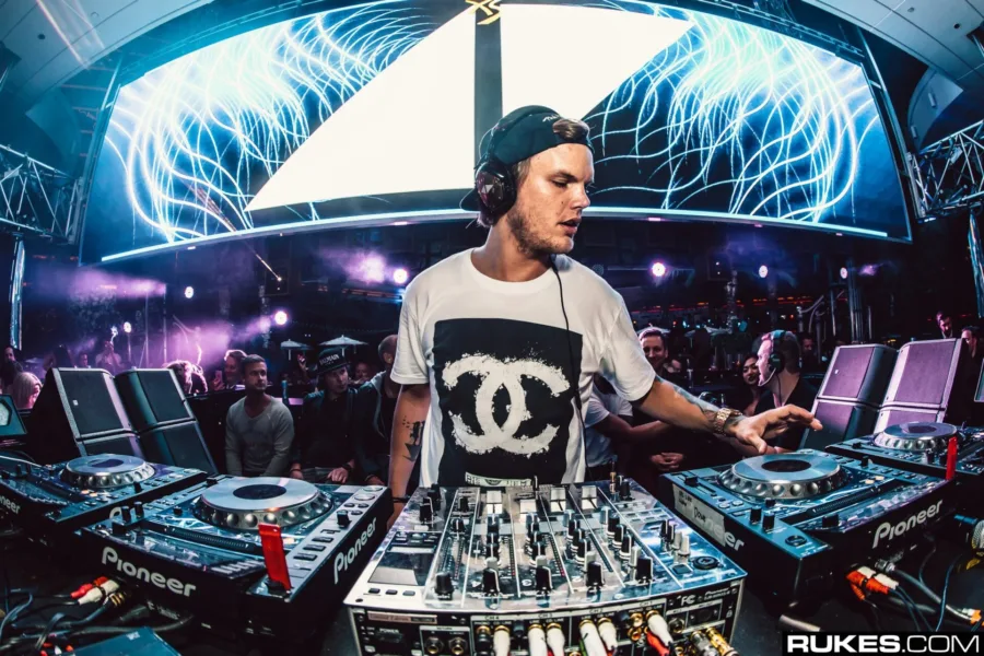 Ο Avicii σε DJ set με εξοπλισμό Pioneer και φόντο μια μεγάλη οθόνη με γραφικά.