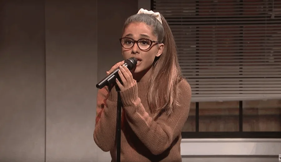 Η Ariana Grande με γυαλιά και καφέ πουλόβερ τραγουδάει σε μικρόφωνο στο Saturday Night Live.