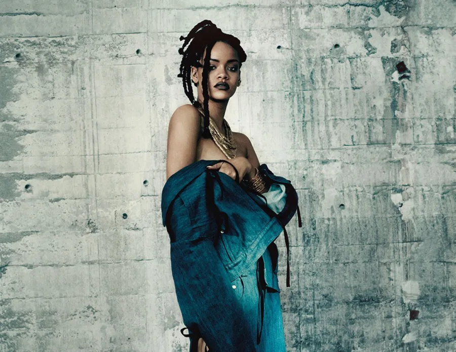 Η Rihanna ποζάρει με denim τζάκετ και dreadlocks μπροστά από τσιμεντένιο τοίχο.