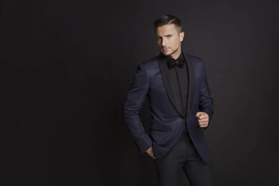 Ο Sergey Lazarev ποζάρει με σκούρο μπλε σμόκιν και μαύρο παπιγιόν σε μαύρο φόντο.