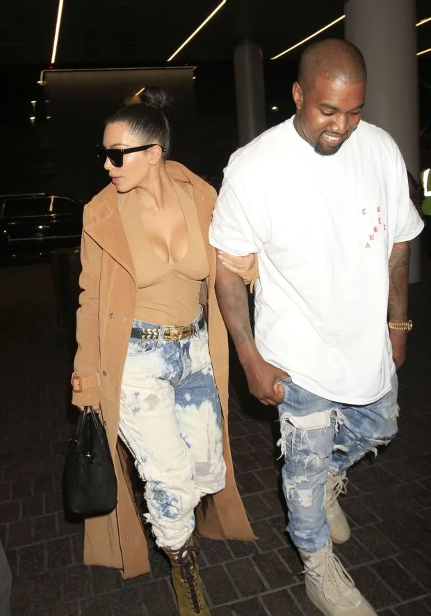 Kim Kardashian & Kanye West are seen flying out of Los Angeles arm-in-arm as Kim shows off her unbelievable assets in a low-cut blouse. Kardashian was seen in torn jeans, a low cut blouse, a trench coat, sunglasses, boots & her hair in a bun. Kanye was