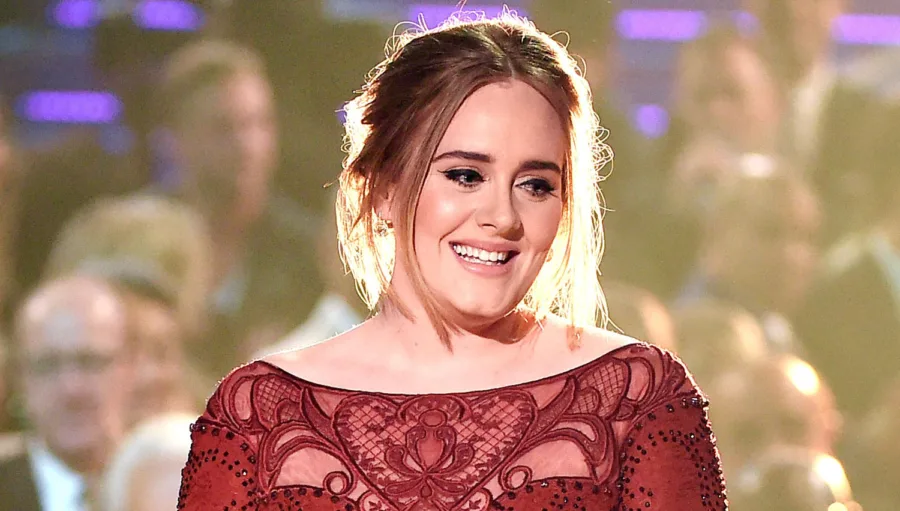 Η Adele χαμογελάει σε εκδήλωση φορώντας ένα μπορντό δαντελωτό φόρεμα.