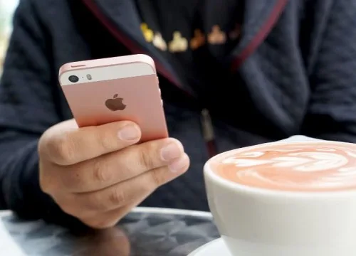 Κοντινό πλάνο ενός ροζ iPhone στα χέρια ενός ατόμου δίπλα σε ένα φλιτζάνι καφέ.