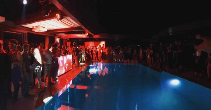 Πλήθος κόσμου σε πάρτι δίπλα σε πισίνα με κόκκινο και μπλε φωτισμό.