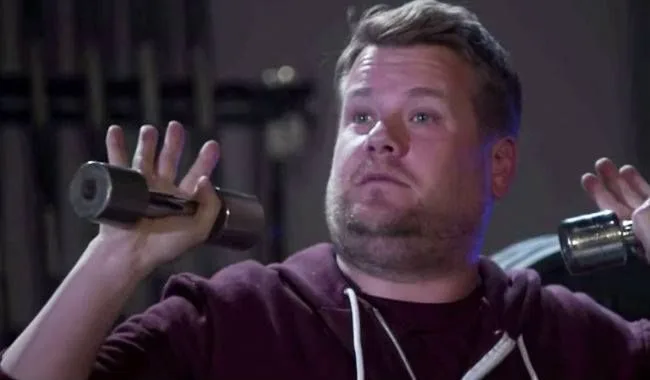 Ο James Corden κρατάει βαράκια σε γυμναστήριο.