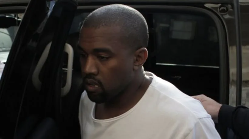 Ο Kanye West με λευκό μπλουζάκι δίπλα σε ένα αυτοκίνητο.