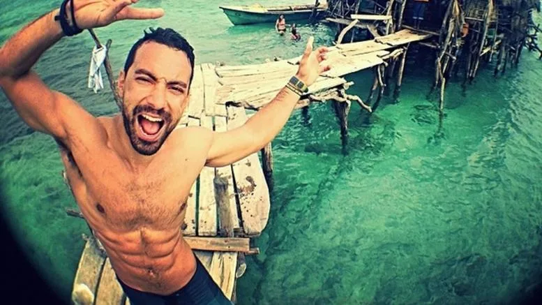 Ο Σάκης Τανιμανίδης ποζάρει χαμογελαστός σε ξύλινη προβλήτα πάνω από γαλαζοπράσινα νερά.