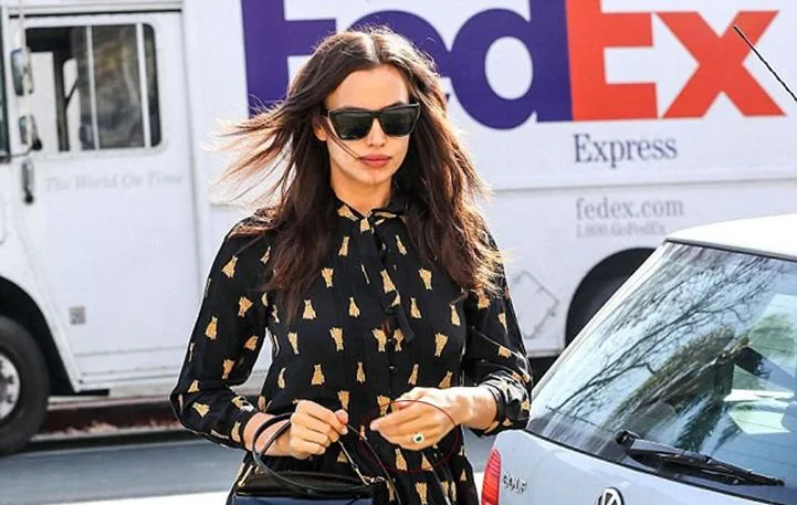 Η Irina Shayk περπατά στο δρόμο φορώντας μαύρο φόρεμα με σχέδια και γυαλιά ηλίου.