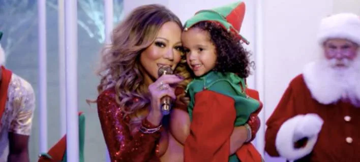 Η Mariah Carey με χριστουγεννιάτικη αμφίεση κρατάει ένα παιδί ντυμένο ξωτικό.