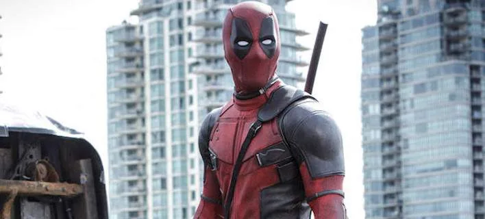 Ο Deadpool με την κόκκινη στολή του σε αστικό τοπίο με κτίρια στο φόντο.
