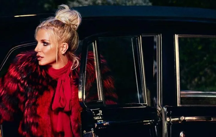 Η Britney Spears με κόκκινη γούνα δίπλα σε μαύρο αυτοκίνητο.