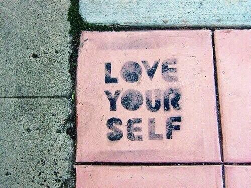Στένσιλ με το μήνυμα LOVE YOUR SELF σε ροζ πλάκα πεζοδρομίου.