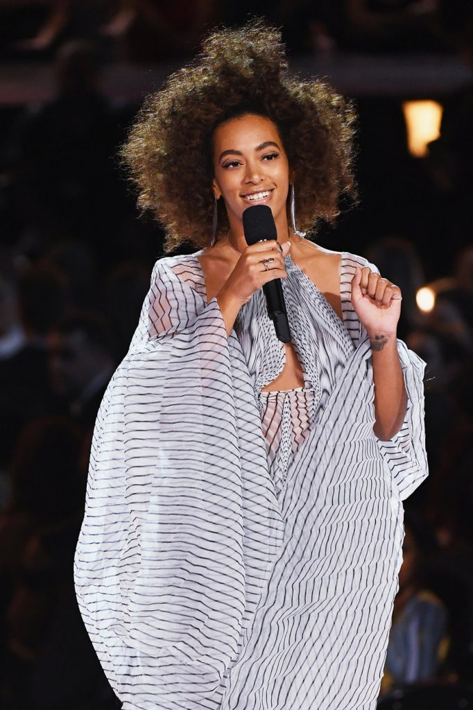 Τραγουδίστρια με afro μαλλιά και ριγέ φόρεμα μιλάει σε μικρόφωνο στα Grammys.