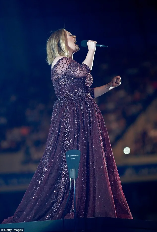 Η Adele τραγουδάει στη σκηνή φορώντας ένα μακρύ μπορντό φόρεμα με παγιέτες.