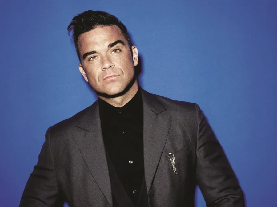 Ο Robbie Williams με γκρι κοστούμι και μαύρο πουκάμισο σε μπλε φόντο.