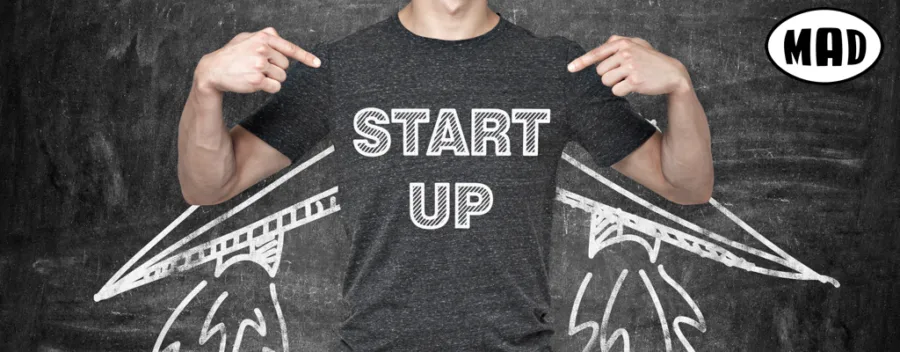 Άνδρας με t-shirt που γράφει START UP δείχνει το στήθος του μπροστά από σχέδιο πύραυλου σε μαυροπίνακα.