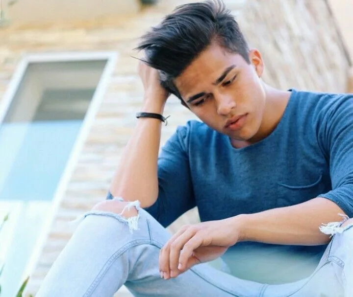 Ο Alex Aiono κάθεται έξω φορώντας μπλε μπλούζα και σκισμένο τζιν.