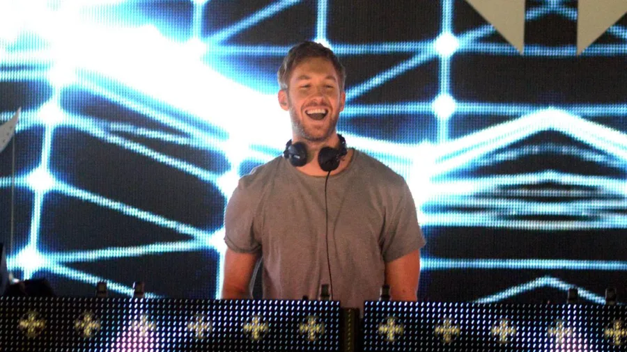 Ο Calvin Harris χαμογελαστός σε DJ set με ακουστικά στον λαιμό.