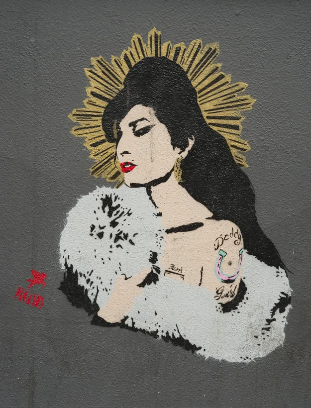 Stencil graffiti της Amy Winehouse με χρυσό φωτοστέφανο σε γκρίζο τοίχο.