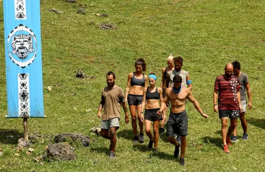 Παίκτες του Survivor περπατούν σε ένα καταπράσινο λιβάδι δίπλα σε μια γαλάζια σημαία.