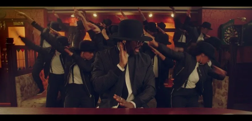 Ο will.i.am με μαύρο καπέλο και ριγέ σακάκι χορεύει μαζί με ομάδα χορευτών σε σκηνικό που θυμίζει παμπ.