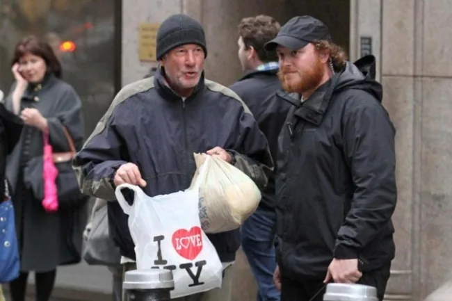 Ο Richard Gere με σκούφο και μπουφάν κρατάει πλαστικές σακούλες στο δρόμο.