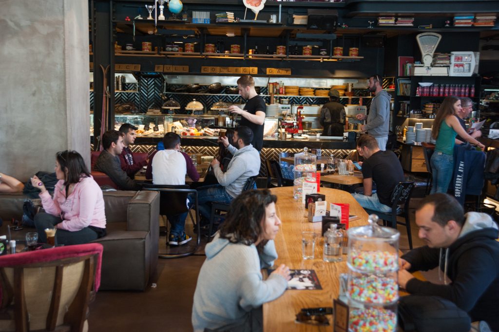 Εσωτερικό του all-day eatery Gazi College με κόσμο να κάθεται σε τραπέζια και μπάρα.