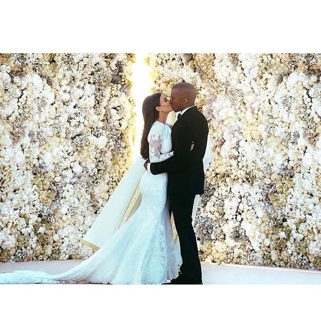 Ο Kanye West και η Kim Kardashian φιλιούνται στον γάμο τους μπροστά από έναν τοίχο με λουλούδια.