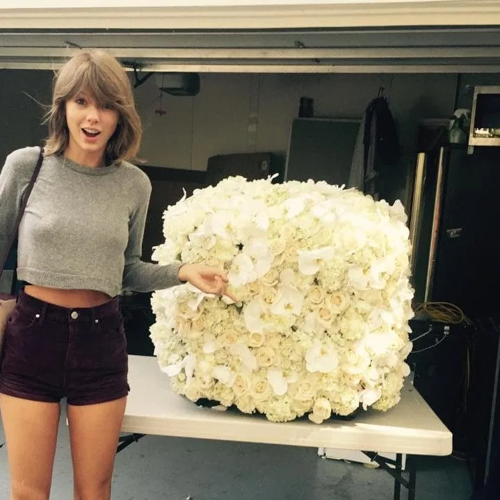 Η Taylor Swift δίπλα σε μια τεράστια σύνθεση από λευκά λουλούδια.