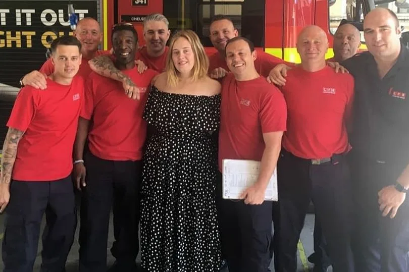 Η Adele ποζάρει ανάμεσα σε ομάδα πυροσβεστών μπροστά από ένα πυροσβεστικό όχημα.