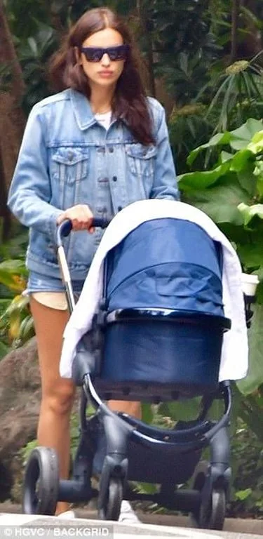 Η Irina Shayk με τζιν τζάκετ και γυαλιά ηλίου σπρώχνει ένα μπλε καρότσι μωρού.