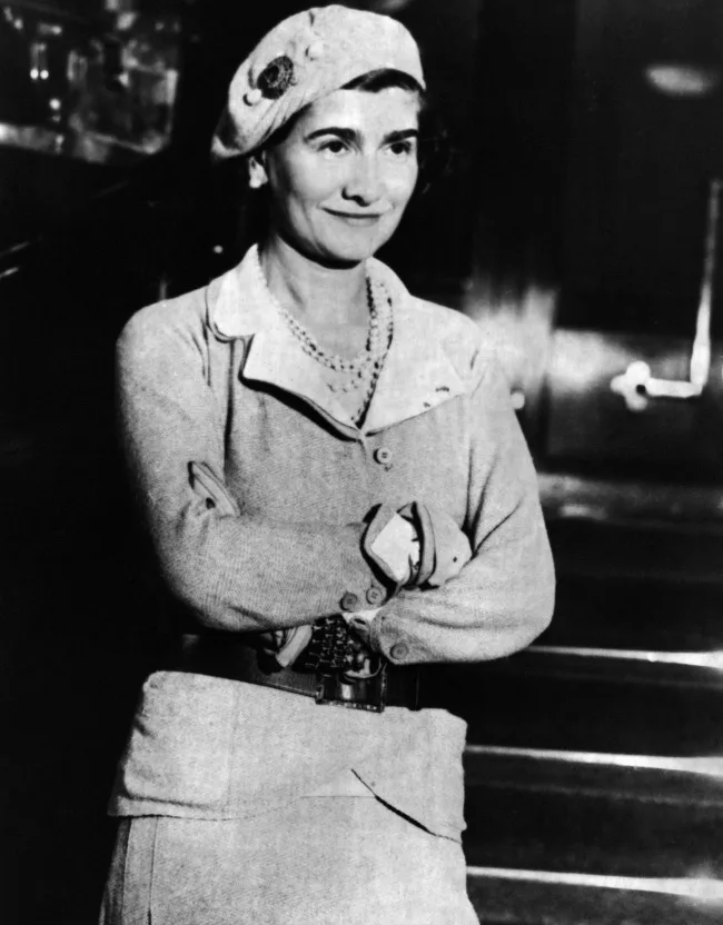Ασπρόμαυρη φωτογραφία της Coco Chanel με μπερέ και πέρλες.