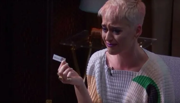 Η Katy Perry με κοντό ξανθό μαλλί κρατάει ένα χαρτάκι και κλαίει.