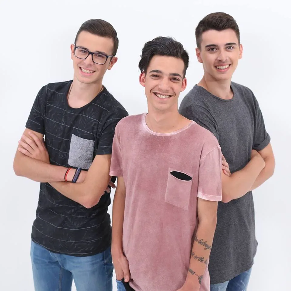 Τρία μέλη ενός boy band ποζάρουν χαμογελαστά σε λευκό φόντο.