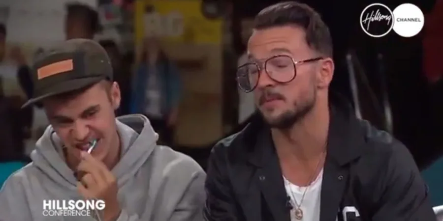 Ο Justin Bieber βουρτσίζει τα δόντια του κατά τη διάρκεια συνέντευξης δίπλα σε έναν άνδρα με γυαλιά.