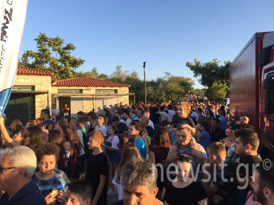Πλήθος κόσμου συγκεντρωμένο σε εξωτερικό χώρο με κτίρια και δέντρα στο βάθος.